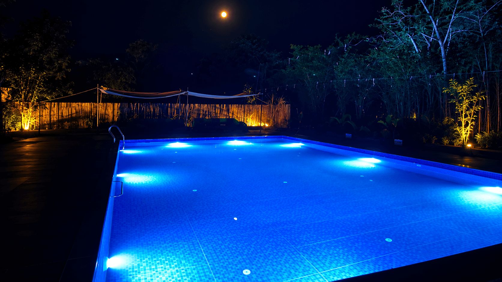Luxury swimming pool at Avani Tiger Resort illuminated with blue underwater lights at night, surrounded by lush trees and warm outdoor lighting.